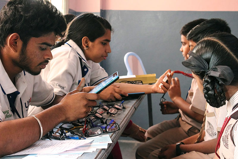 A male ophthalmic technician is holding a mobile phone with the Peek app on and a female ophthalmic technician holds a pair of spectacles. They are sitting opposite three children, at a table with many pairs of spectacles on it.