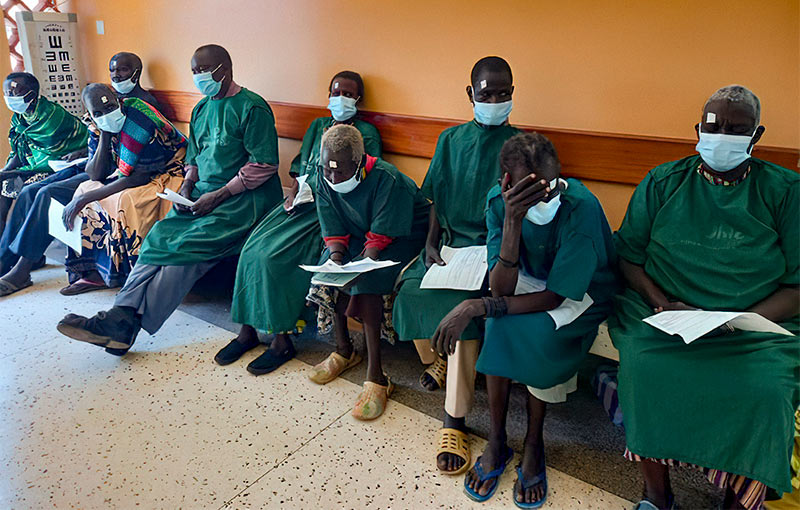 Nine patients sitting donw on a bench in a waiting area. Each of them are holding papers and wearing face masks and dark green surgical gowns.