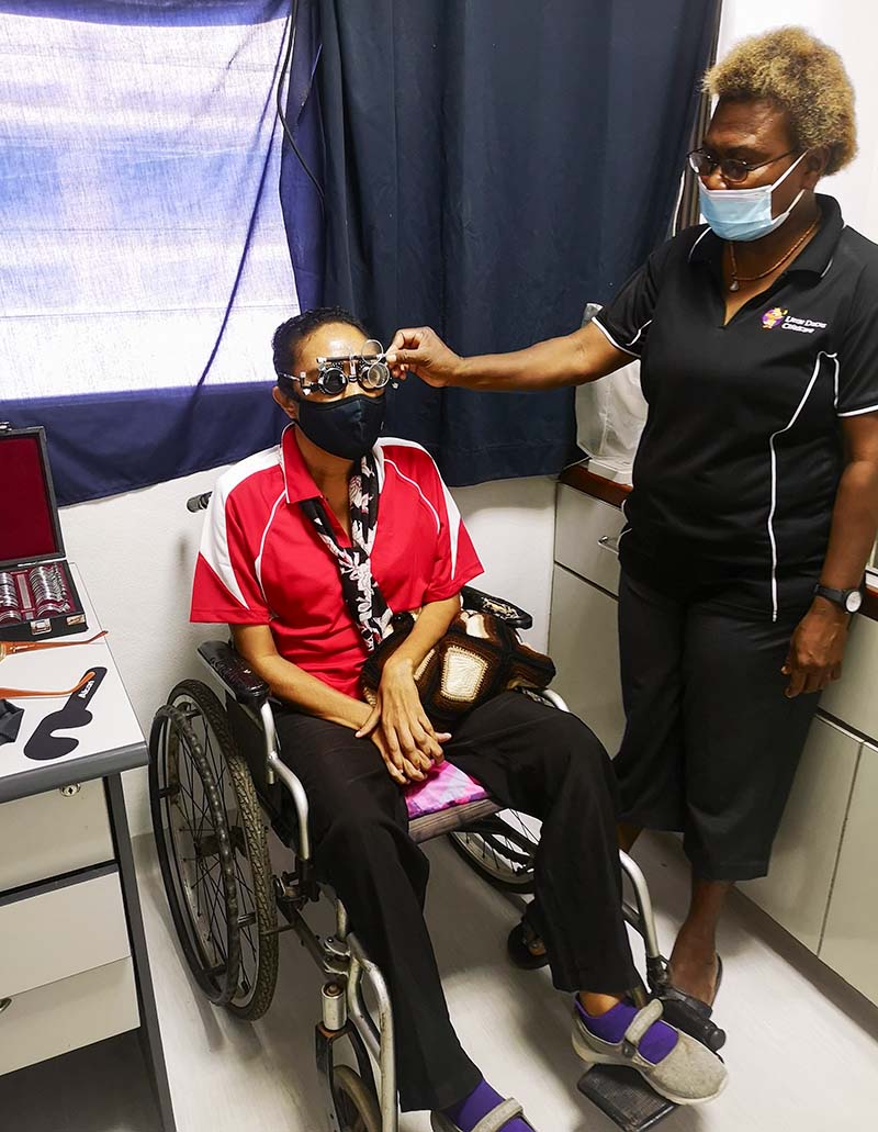 A female carer holds refraction spectacles up to the eyes of a female patient who is sitting in a wheelchair.