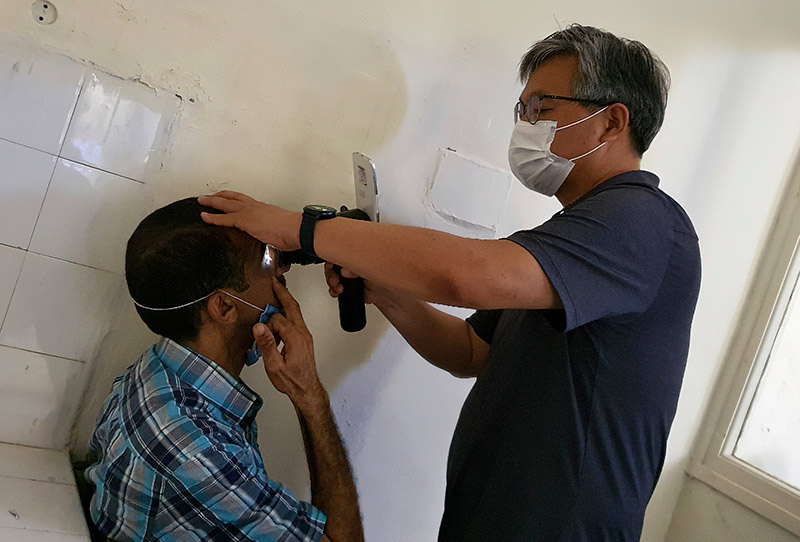 A male ophthalmologist examines the eye of a male patient who is sitting down.