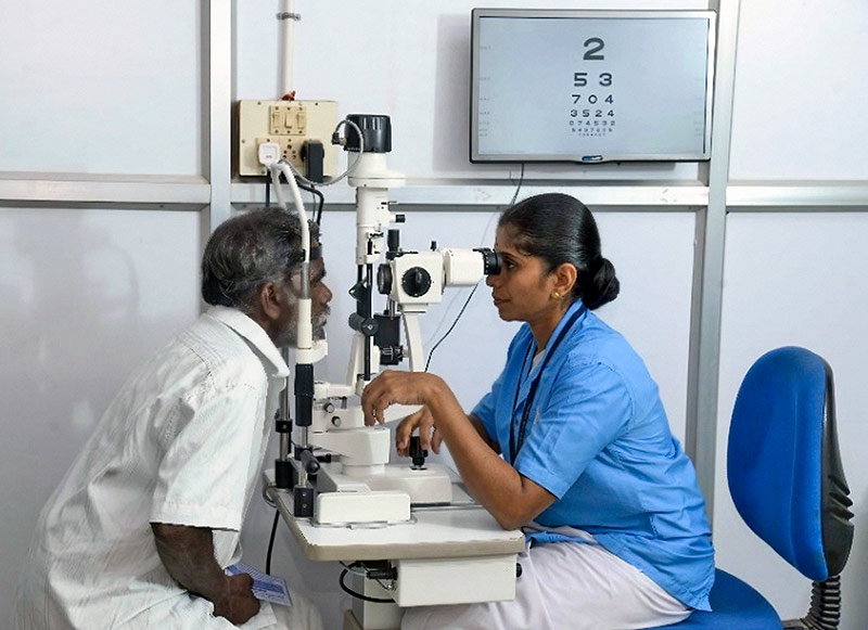 A female vision technician examines a man's eyes in clinic. Both are seated at the slit lamp.