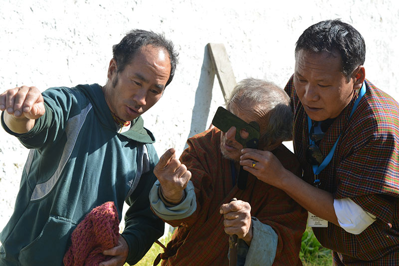 Two eye care workers either side of an elderly man, checking vision while holding a cover over one of his eyes