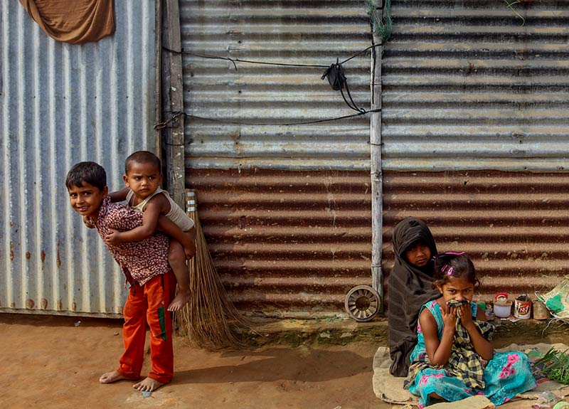 Four children outside a house. One child (boy) is carrying another child (boy) on his back, the other two children (girls) are sitting down on the ground.