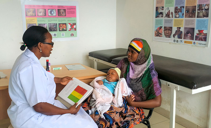 A nurse is sitting across from a mother who is holding her baby in the clinic. The nurse is holding a book open on a page with red, yellow and green sections.
