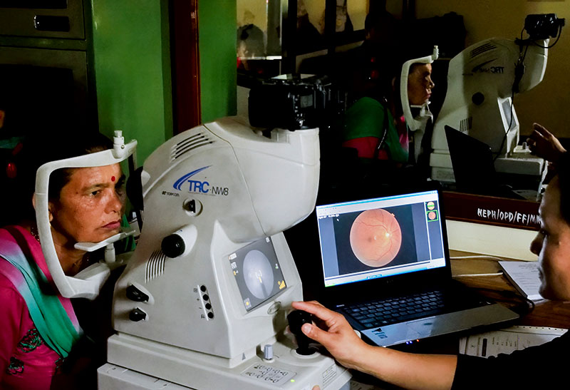 A female patient sits with her head on the chin rest of the machine which is being operated by a member of the eye care team
