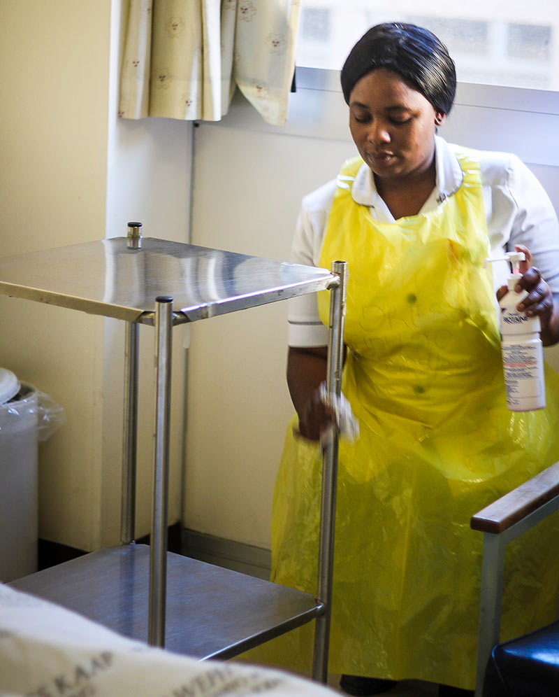 A female hospital staff member holding a pump container of disinfectant, wiping down the legs of a metal trolley