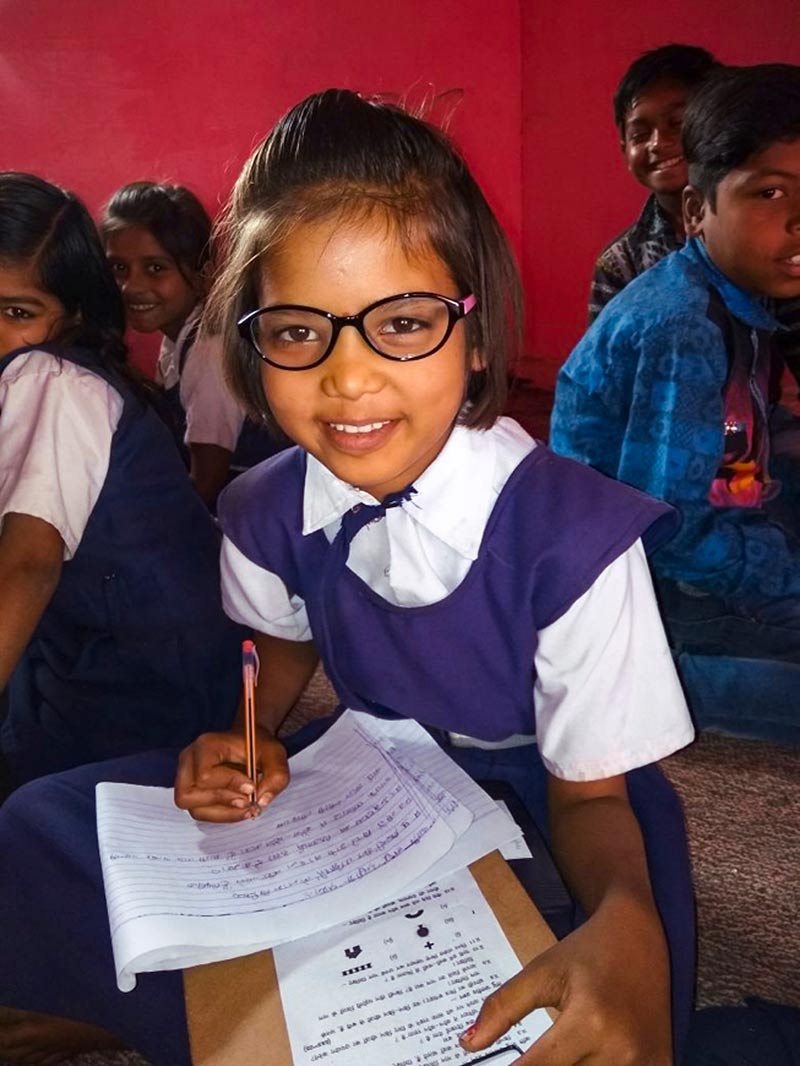 A young school girl wearing spectacles, smiling. She is holding a pen and writing on a lined sheet of paper with her left hand. She is holding a clipboard with a question sheet in her right hand.