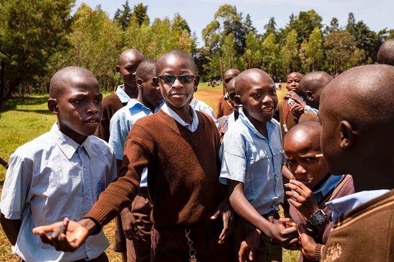 a group of school boys, one of whom is wearing spectacles are standing outside in the sunshine
