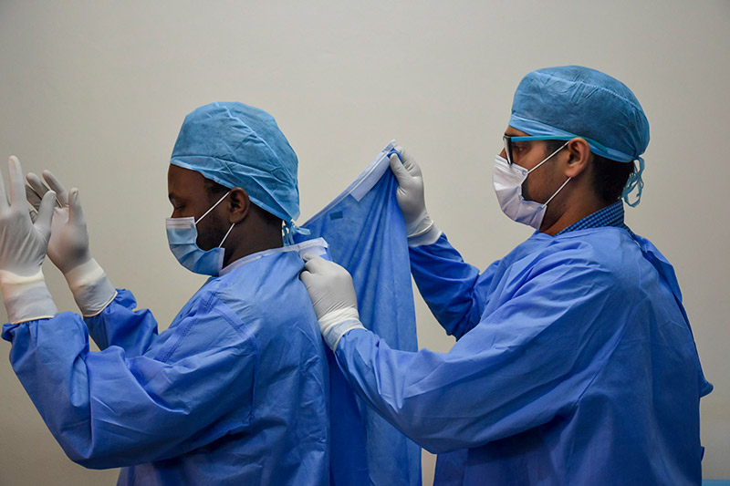 A health care worker wearing a blue gowns, blue surgical caps, surgical mask and gloves helping a colleague to fasten the back of their gown