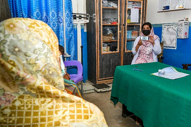 A female health visitor holds a smartphone up, with the Peek visual acuity app on it, in front of a female patient during a visual acuity test in a clinic room. App