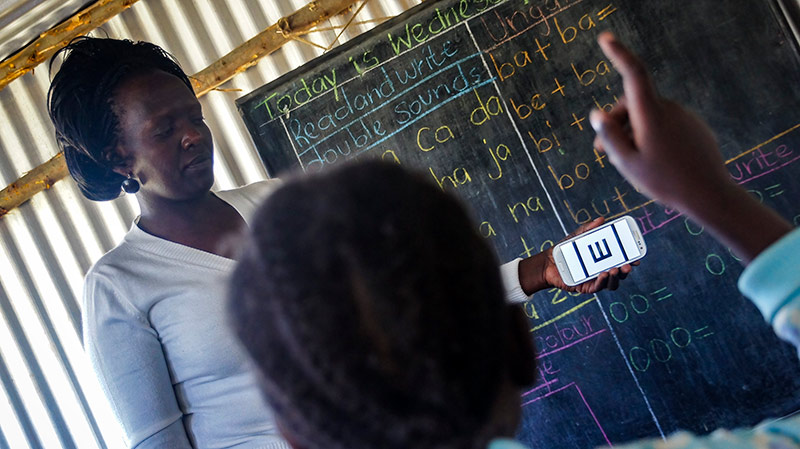 A female teacher holds a mobile phone up at the front of a classroom. The phone display shows the Peek visual acuity app with the tumbling E chart and a child is pointing to indicate the direction of the 'E'