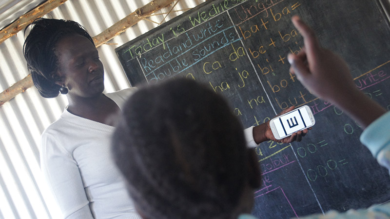 A female teacher in Kenya holding up a smartphone in front of the blackboard, testing vision of a female student
