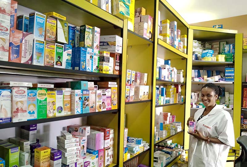 the inside of a pharmacy shop. Many shelves filled with drugs. A smiling staff member, wearing a white coat stands to the side.