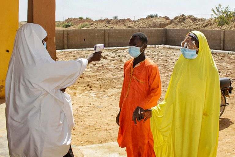 A female health worker points an infrared thermometer towards the forehead of a female patient arriving at the clinic with her male relative escort