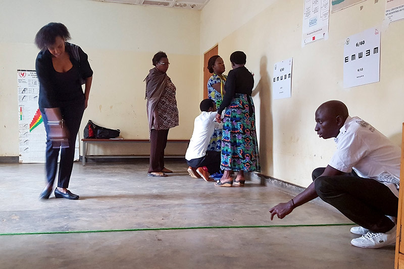 Five eye care workers preparing a room for visual acuity testing. A male eye care worker squats down while holding some green tape across the floor and a female eye care worker points to the floor indicating where the tape should be placed. The other eye care workers are behind them, talking and fixing tumbling E visual acuity charts to the wall