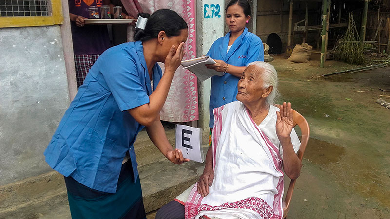 Two feamle eye care workers explain to an elderly female patient who is sititing down outside how to cover one of her eyes ready to have her vision tested with a tumbling E chart