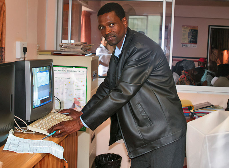 A male member of the hospital staff is standing up at the desk in front of a desktop computer monitor and keyboard, entering details from a sheet of paper.