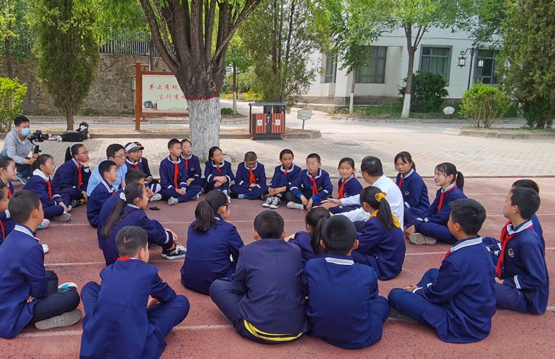 A group of school children wearing dark blue uniforms, sittign outside in a circle under a tree with their teacher